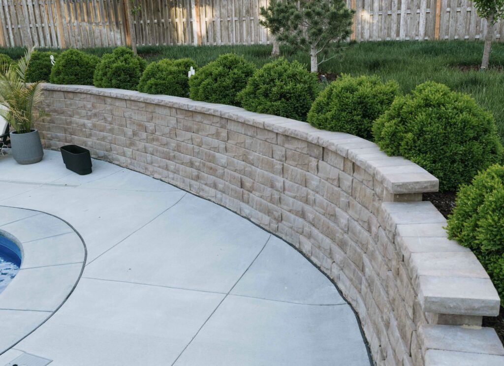 Curved block retaining wall with stone coping lining concrete pool patio and evergreen garden bed