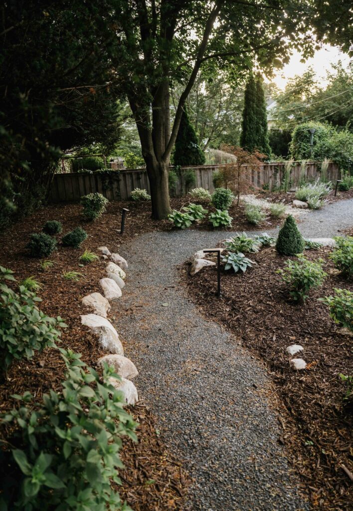 Here are high-intent, SEO-optimized alt text options tailored for this garden walkway photo. Select the variation that best matches your target focus or local market. Primary SEO Alt Text Options High-Intent / Conversion Focused: "Custom gravel walkway through mulched garden bed with stone rock edging and low-voltage landscape lighting