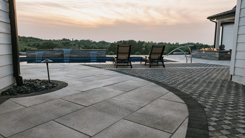 Custom stone paver pool deck with dark accent border, sheer descent pool waterfalls, and lounge seating at sunset