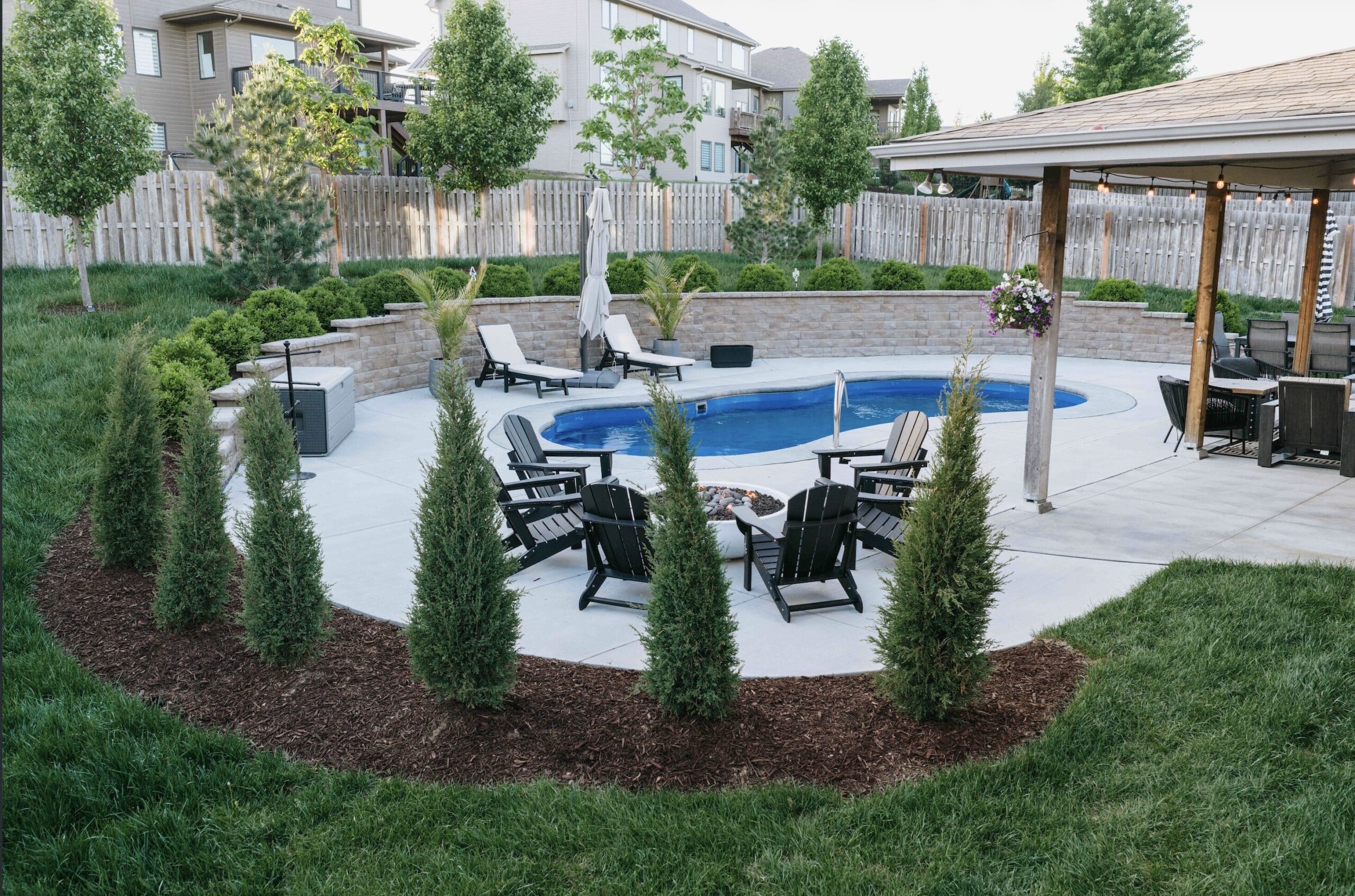 Circular paver fire pit lounge with black metal chairs framed by arborvitae privacy trees