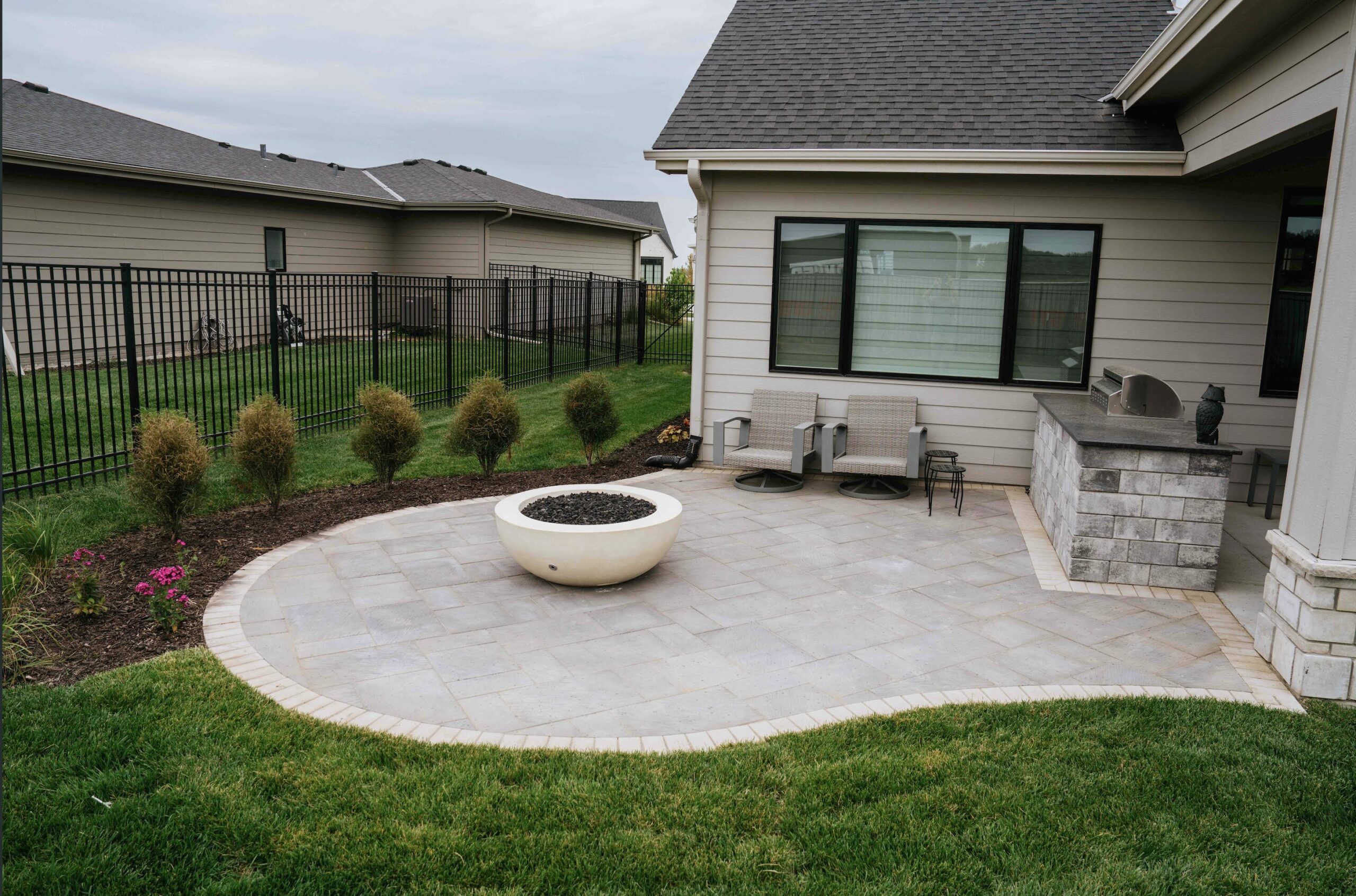 Backyard paver patio featuring a modern fire pit, landscaped lawn, ornamental shrubs, and comfortable outdoor seating.