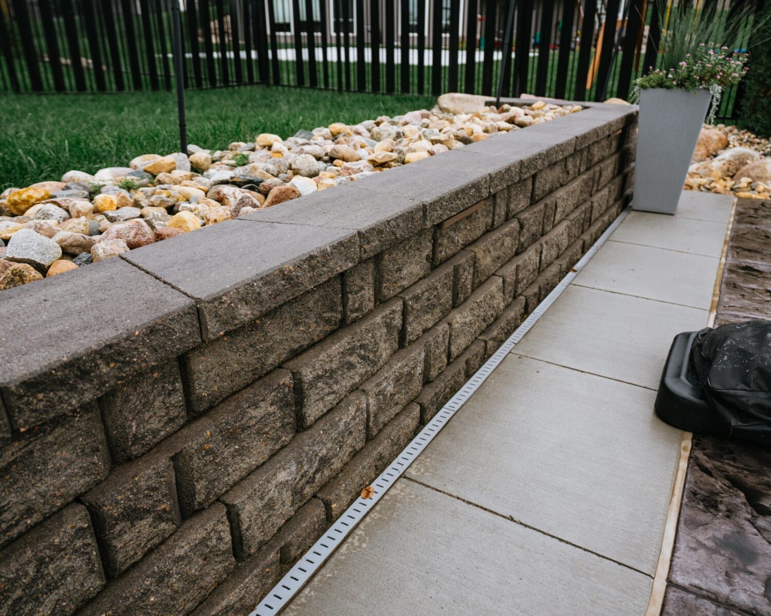 Textured block retaining wall with capstones and river rock drainage channel along patio