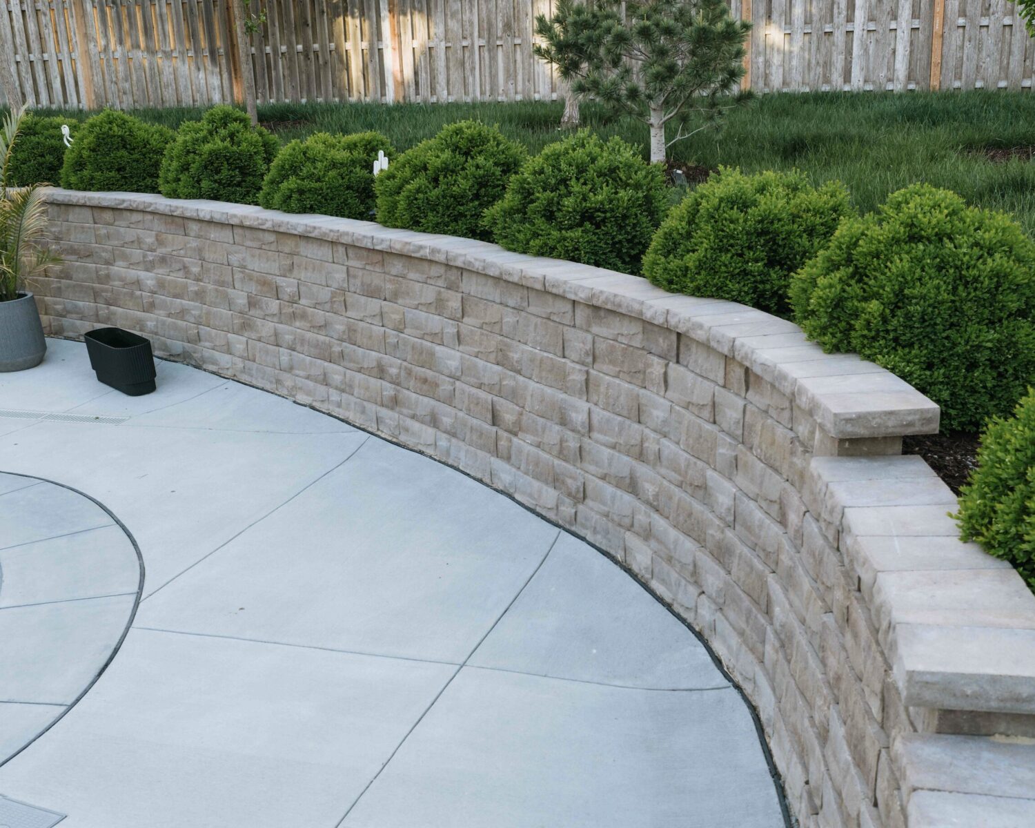 Curved block retaining wall with stone coping lining concrete pool patio and evergreen garden bed