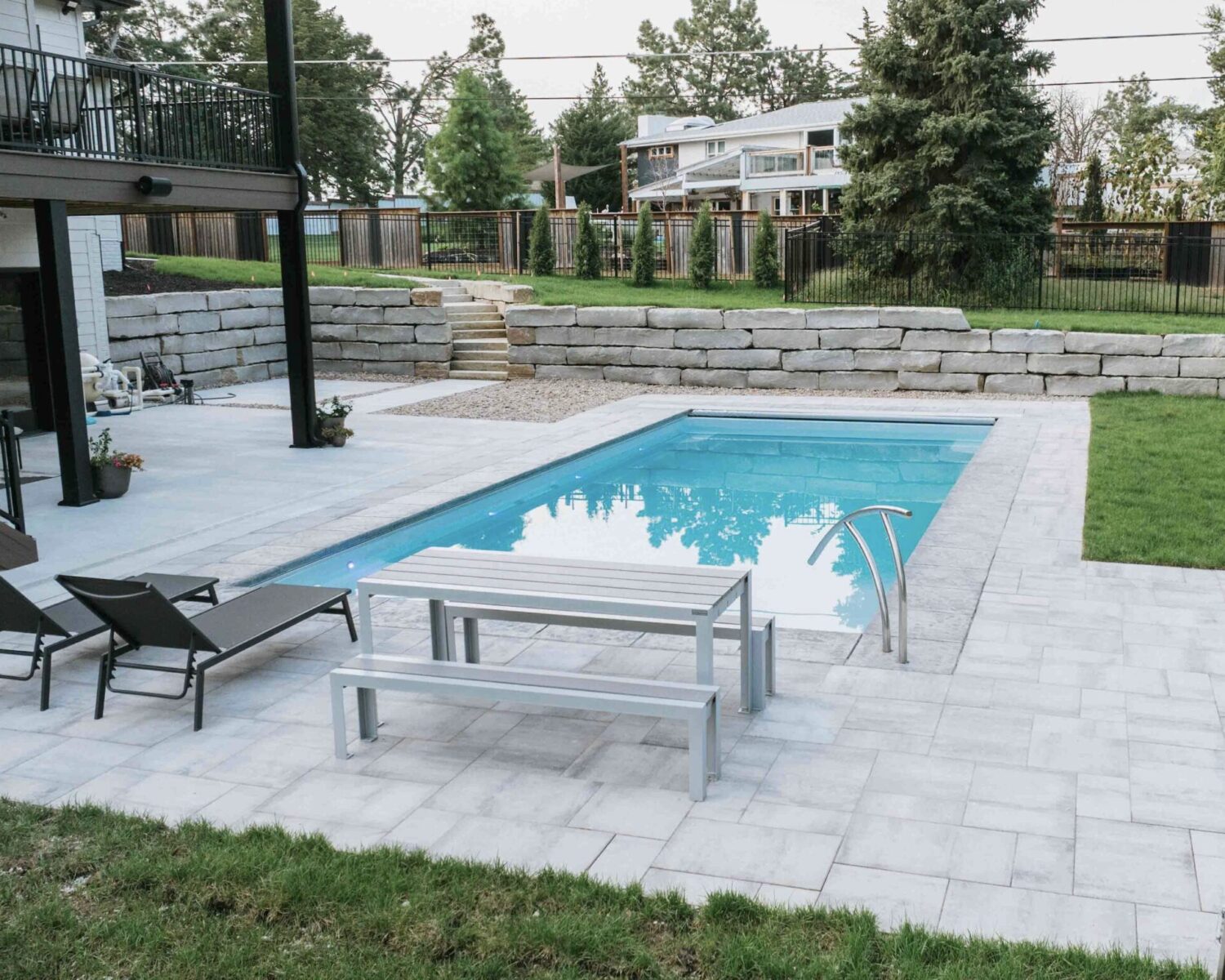 Residential pool with large paver patio, outdoor lounge chairs, landscaped lawn, and stone retaining wall.