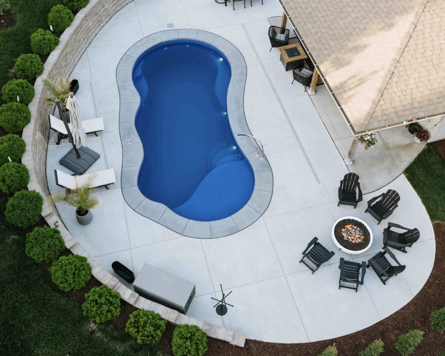 Aerial drone view of custom freeform pool, surrounding concrete patio deck, and outdoor fire pit lounge