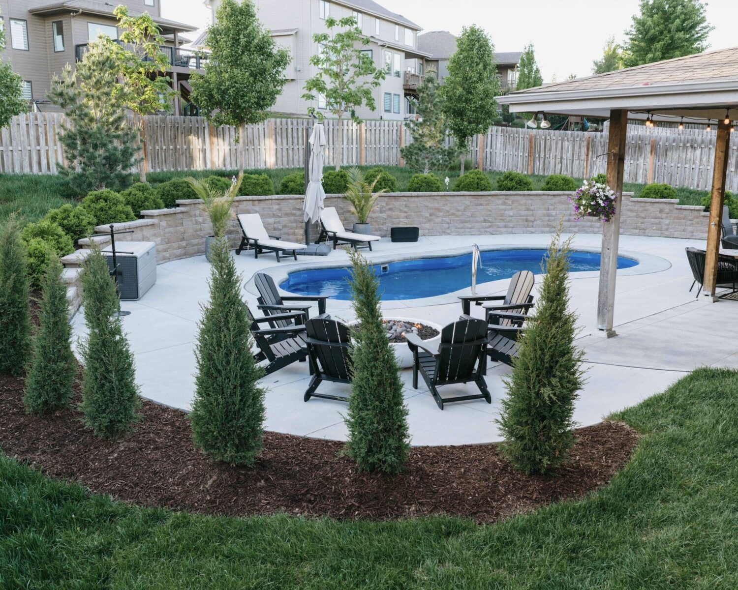 Curved inground pool with privacy tree landscaping, patio lounge chairs, and perimeter mulch beds