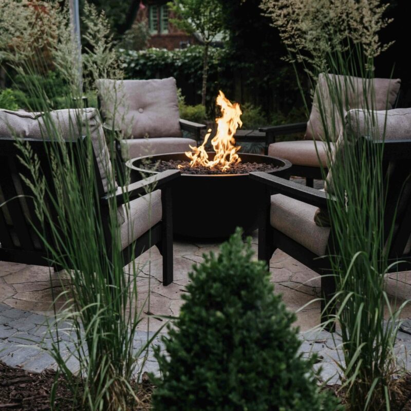 Square gas fire pit table with active flames set in lush backyard garden seating area