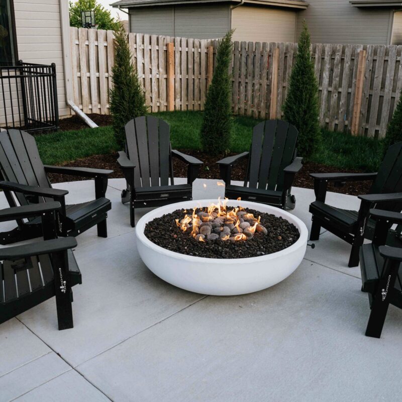Round white concrete gas fire bowl with dark adirondack seating on paver patio