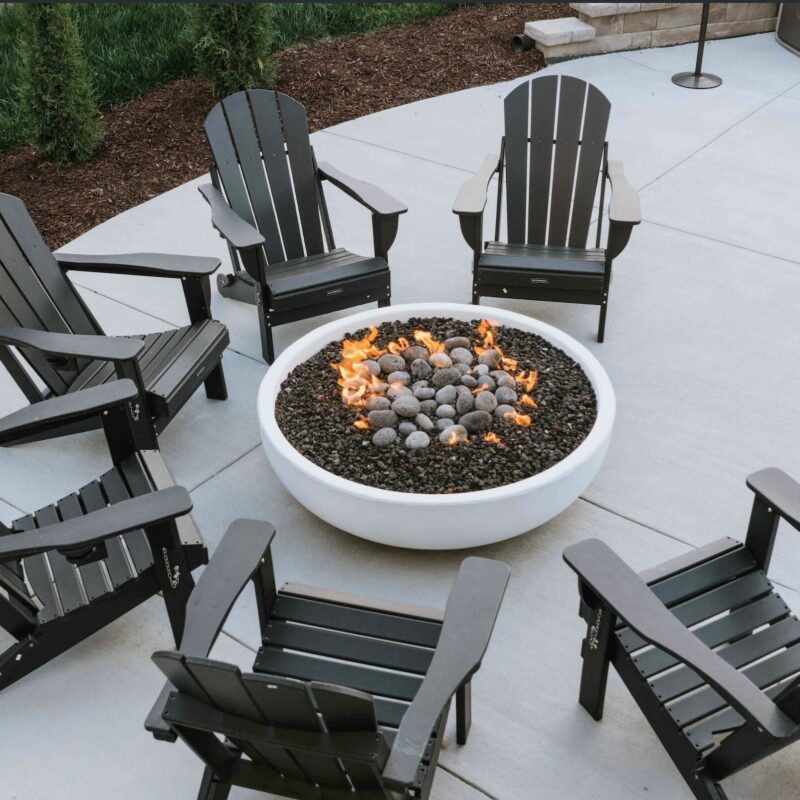 Round white concrete gas fire bowl surrounded by dark adirondack patio chairs