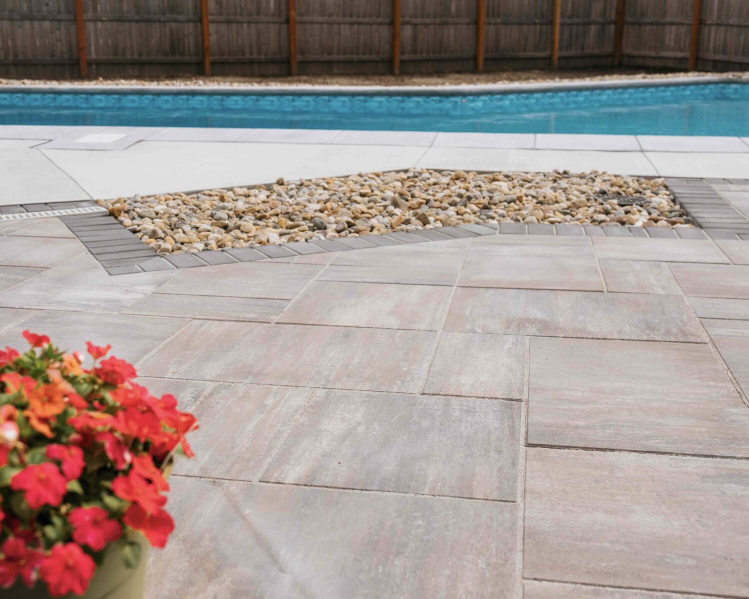 Custom paver pool deck with river rock drainage insert, potted flowers, and privacy fence