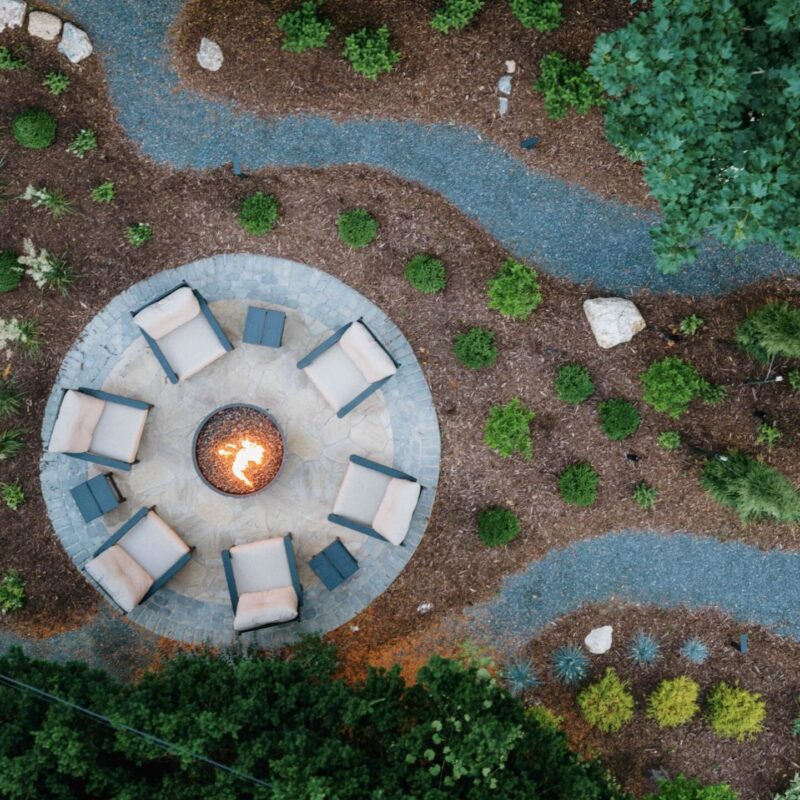 Aerial drone view of round stone patio with fire pit, white chairs, and winding gravel path