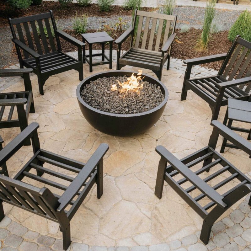 Round black metal fire pit bowl surrounded by dark wooden adirondack chairs on stone patio